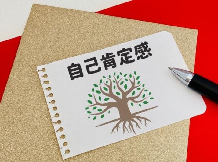 緑色の葉っぱがたくさんついている樹木の絵と自己肯定感と書かれた紙とボールペンがテーブルの上に置かれている様子